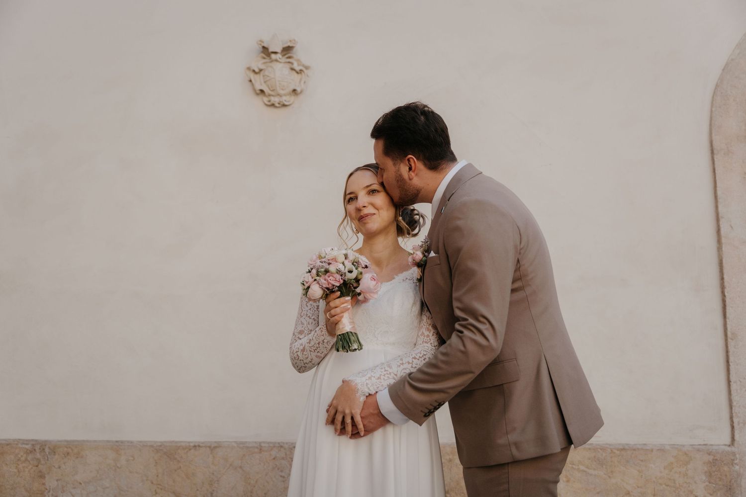 Romantic wedding couple shares tender moments in an elegant outdoor setting with white stone walls.