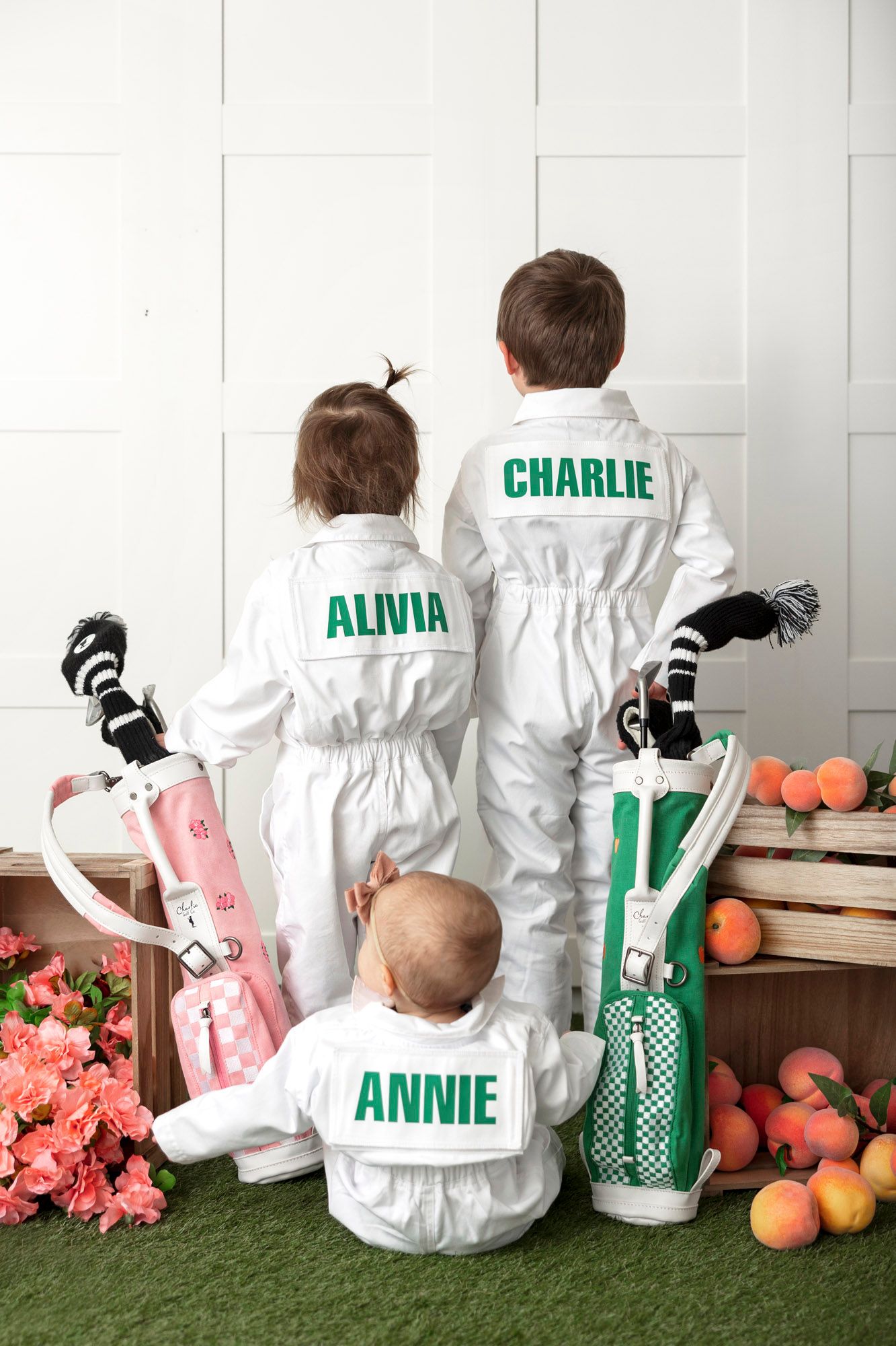 Children in white coveralls with personalized names sit on floor with pumpkins and pink flowers in background.
