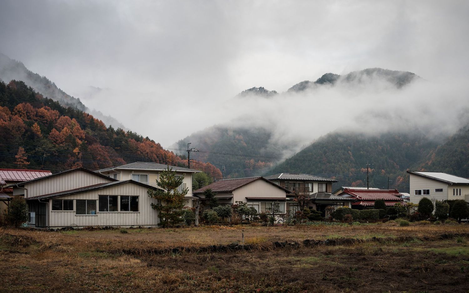 Misty Japanese village with traditional houses nestled among autumn mountains shrouded in low clouds on an overcast day.