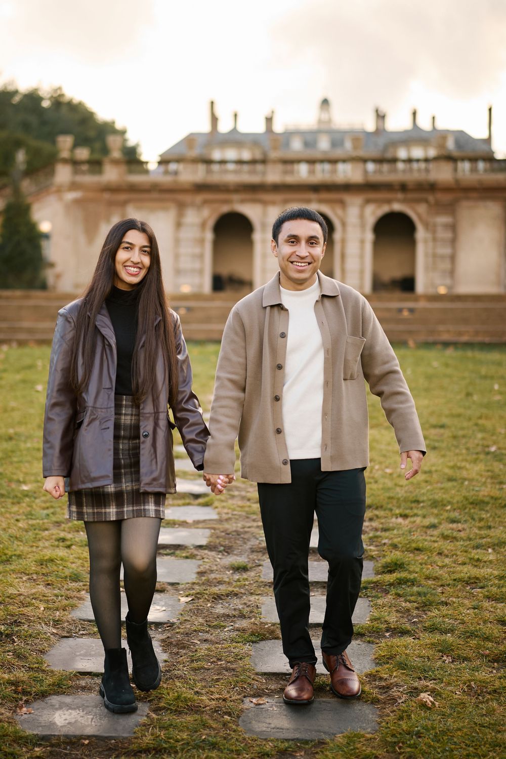 A smiling couple holds hands while walking on a stone path in front of a grand historic building during autumn.