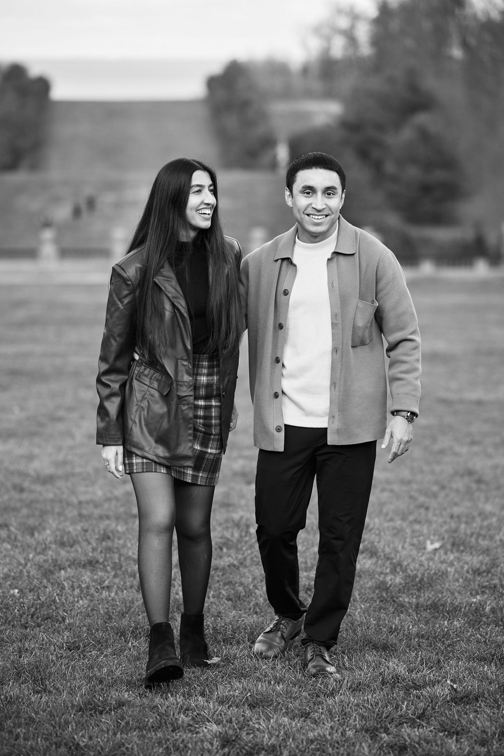 Black and white photo of a smiling couple walking together on a grassy field in stylish autumn outfits.