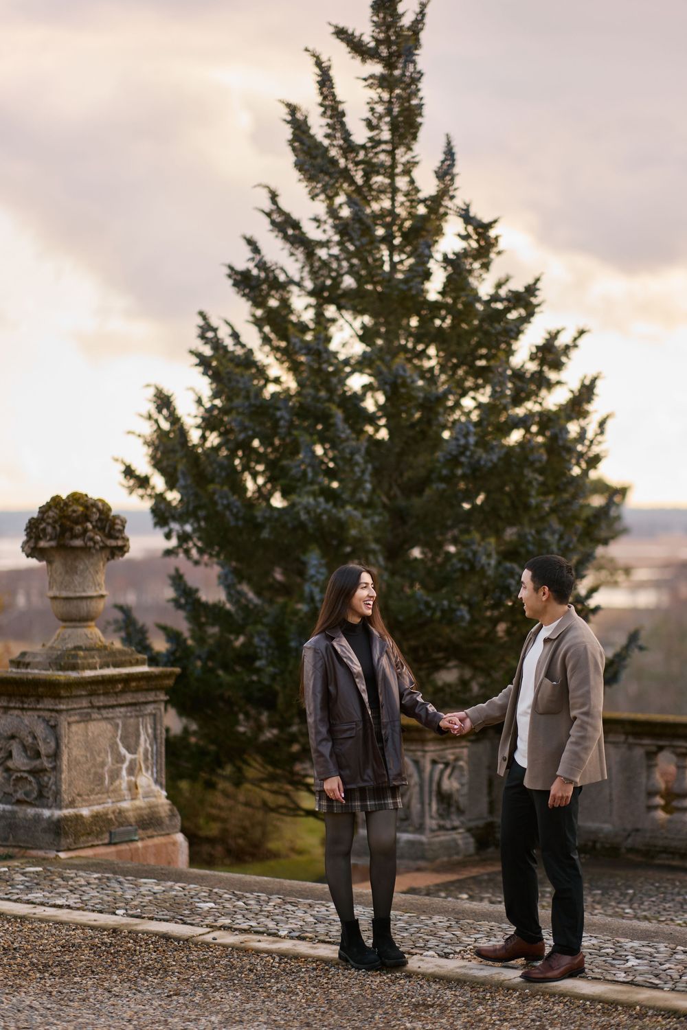Couple holding hands on a stone terrace with a large evergreen tree and dramatic sunset sky backdrop.