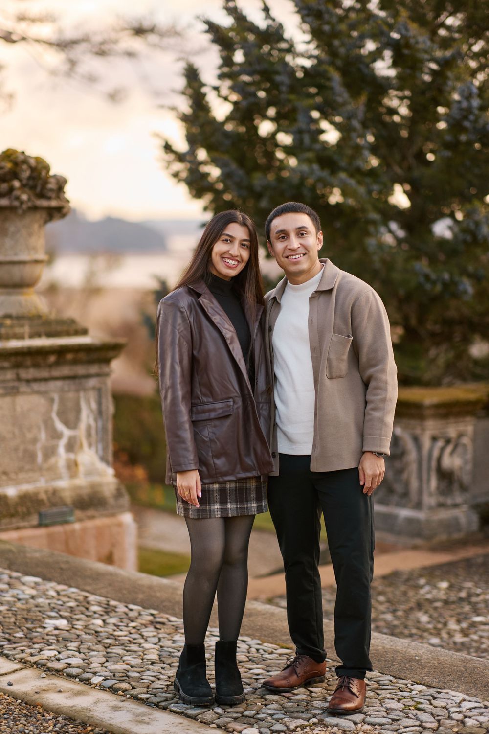 A smiling couple poses together on a cobblestone path in a garden at sunset, wearing casual autumn outfits.