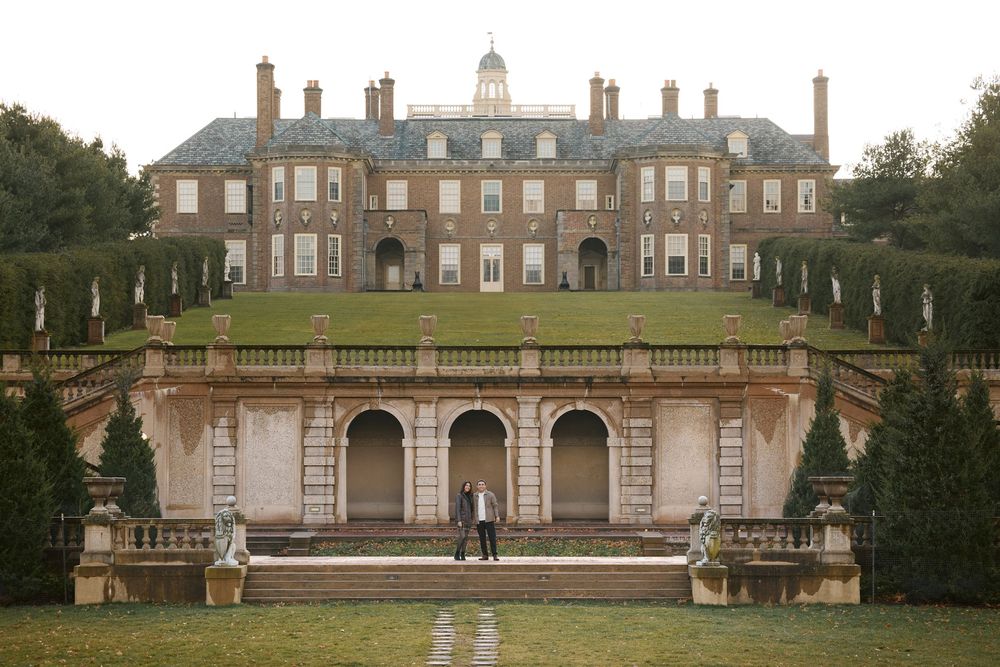 Couple stands before a grand Georgian mansion with terraced gardens, stone balustrades, and manicured lawns at golden hour.