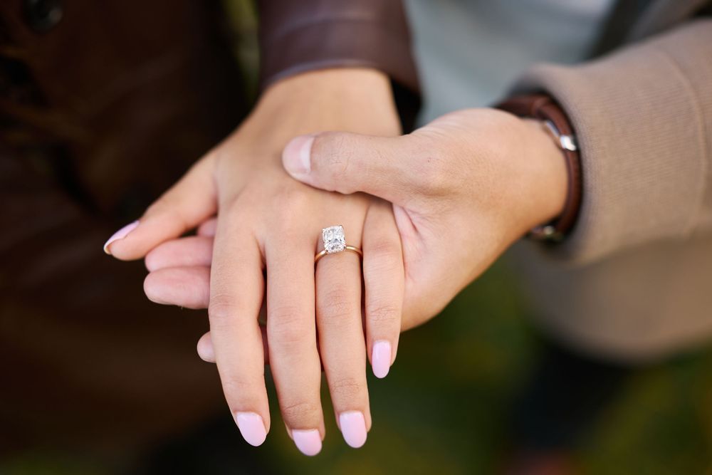 Close-up of a woman's hand displaying a sparkling diamond engagement ring with pink manicured nails resting on a man's hand.