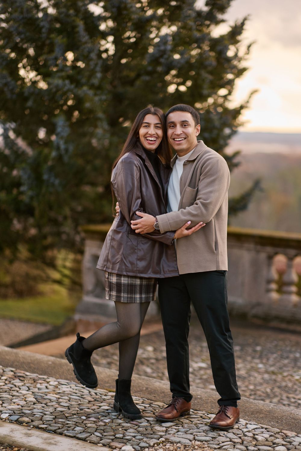 A smiling couple embraces outdoors on a cobblestone path with autumn trees and a scenic landscape at sunset.