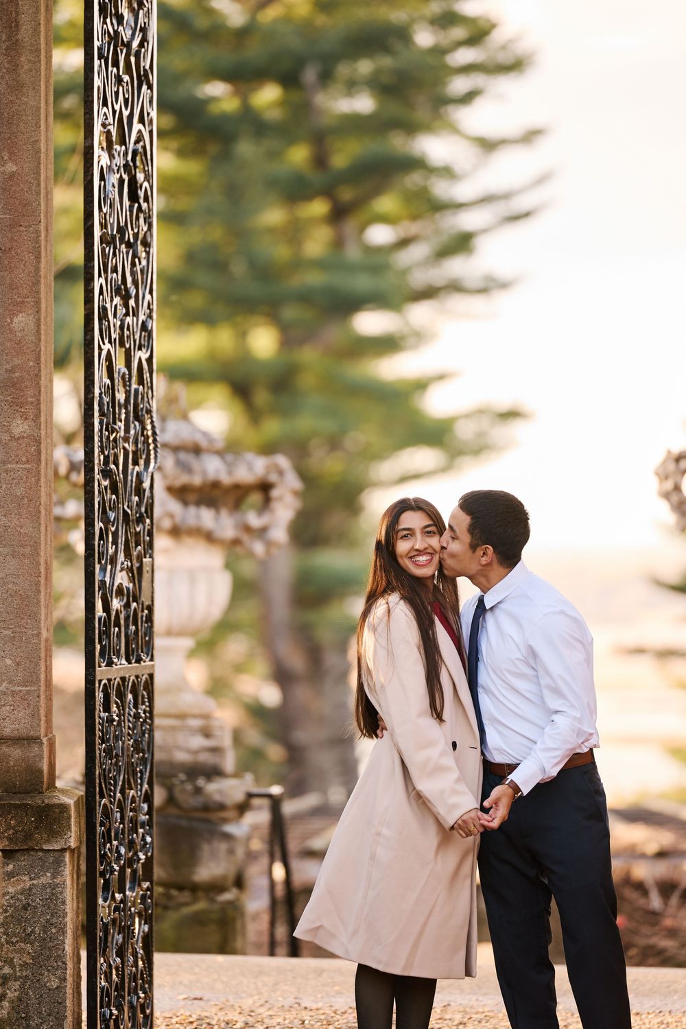 A couple shares a romantic moment by ornate gates, man kissing woman's cheek as she smiles in a beige coat.