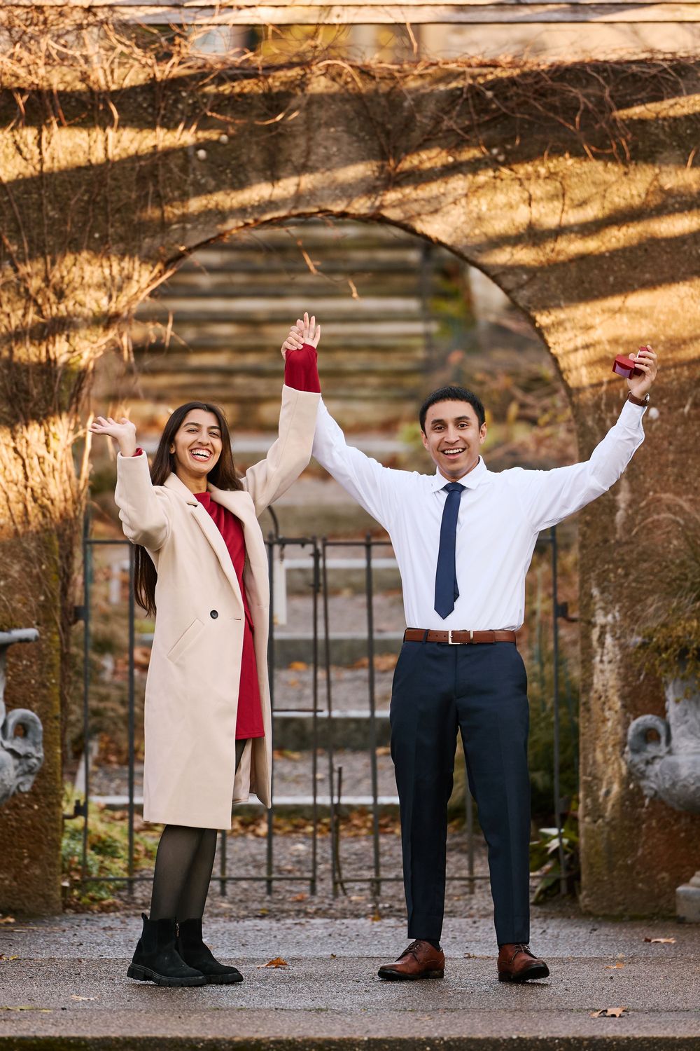 A joyful couple celebrates with raised arms in an autumn garden, woman in beige coat and man in white shirt and tie.