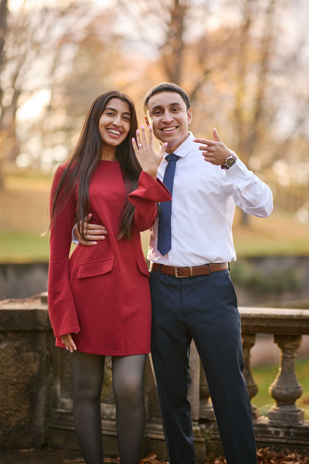 Happy couple posing outdoors in autumn park, woman in red dress showing engagement ring, man in white shirt and blue tie.