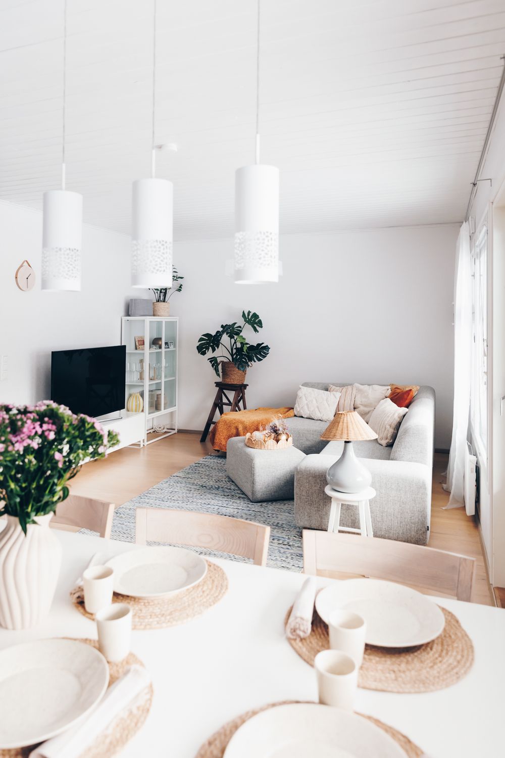 Bright Scandinavian living room with white pendant lamps, grey sofa, dining area with woven placemats and pink flowers.