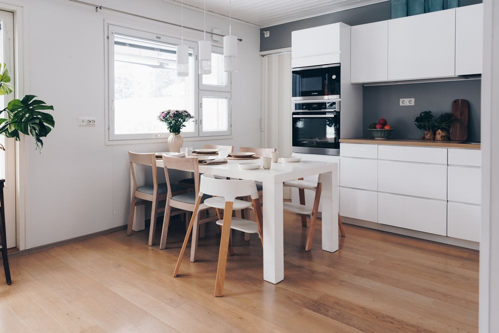 Modern white kitchen with dining area featuring hardwood floors, built-in appliances, and Scandinavian-style furniture.