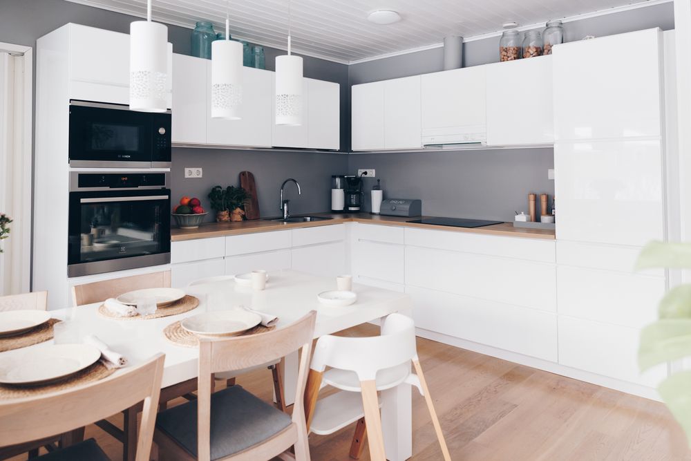Modern white L-shaped kitchen with wooden dining table, white chairs and built-in appliances in a minimalist design.