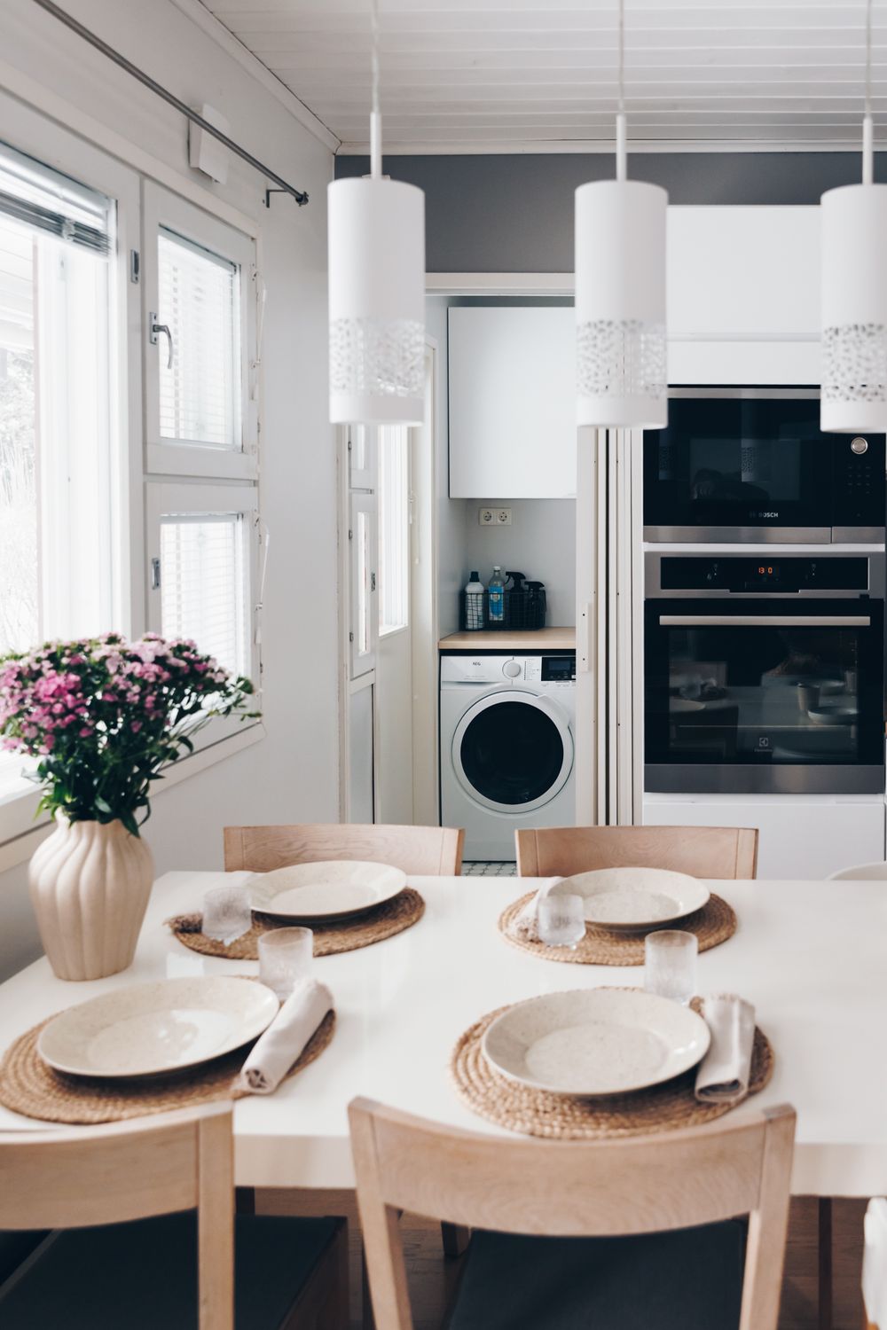 Modern white dining room with wooden table, pendant lights, and minimalist place settings in a Scandinavian style.