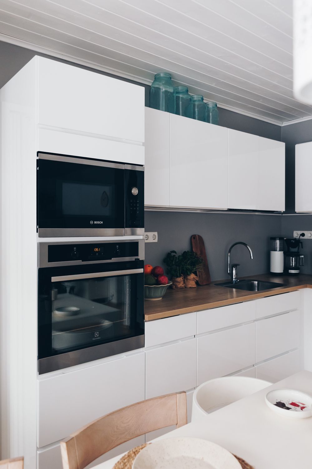 Modern white kitchen with built-in appliances, wooden countertop, and blue glass bottles displayed on top shelf.