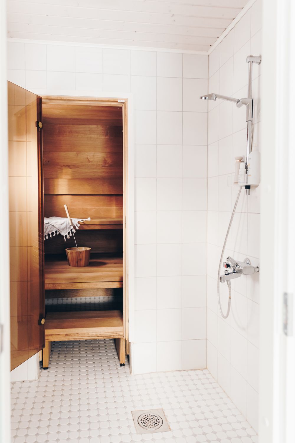 Modern white bathroom interior with wooden sauna benches and chrome shower fixtures on tiled floor.