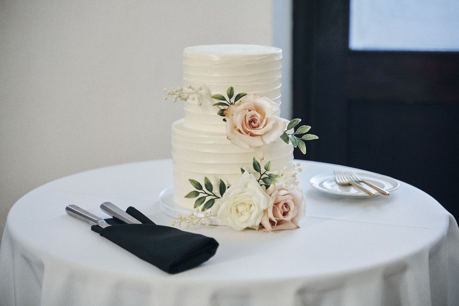 Elegant two-tier white wedding cake decorated with delicate pink roses and greenery sits on a round table with serving utensils.