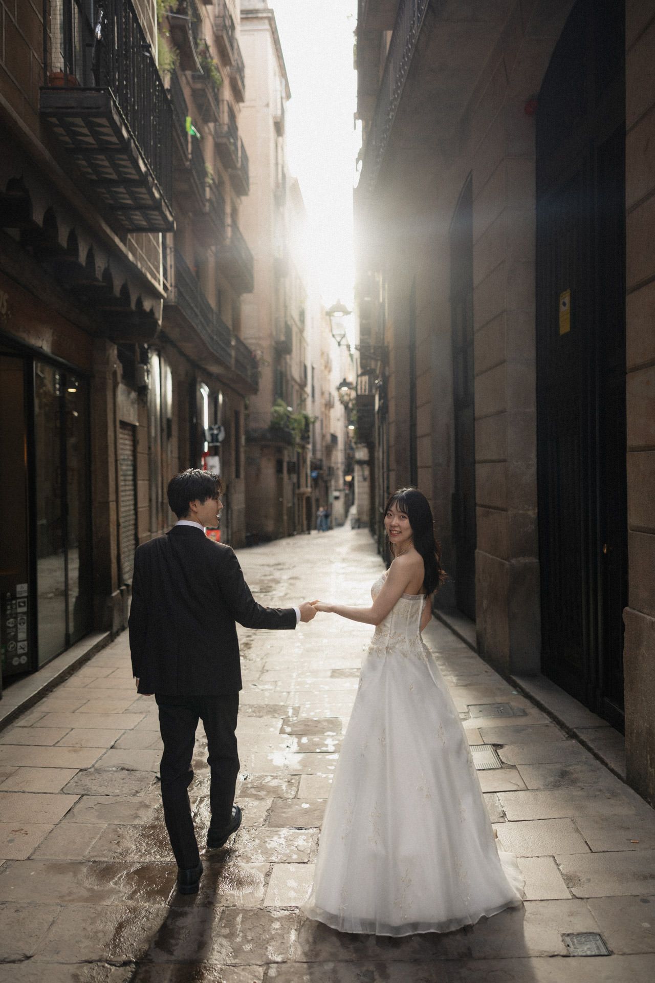 A couple in wedding attire walks through a misty narrow European street with dramatic lighting and historic architecture.