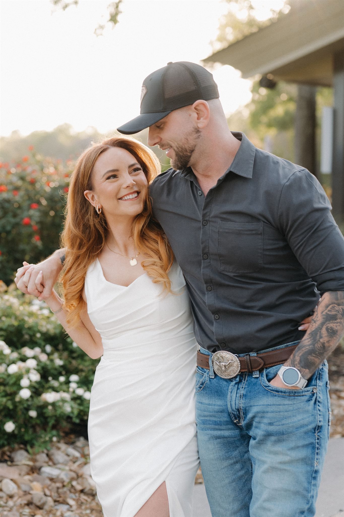 A couple shares intimate moments in a garden setting with white roses and greenery in the background.