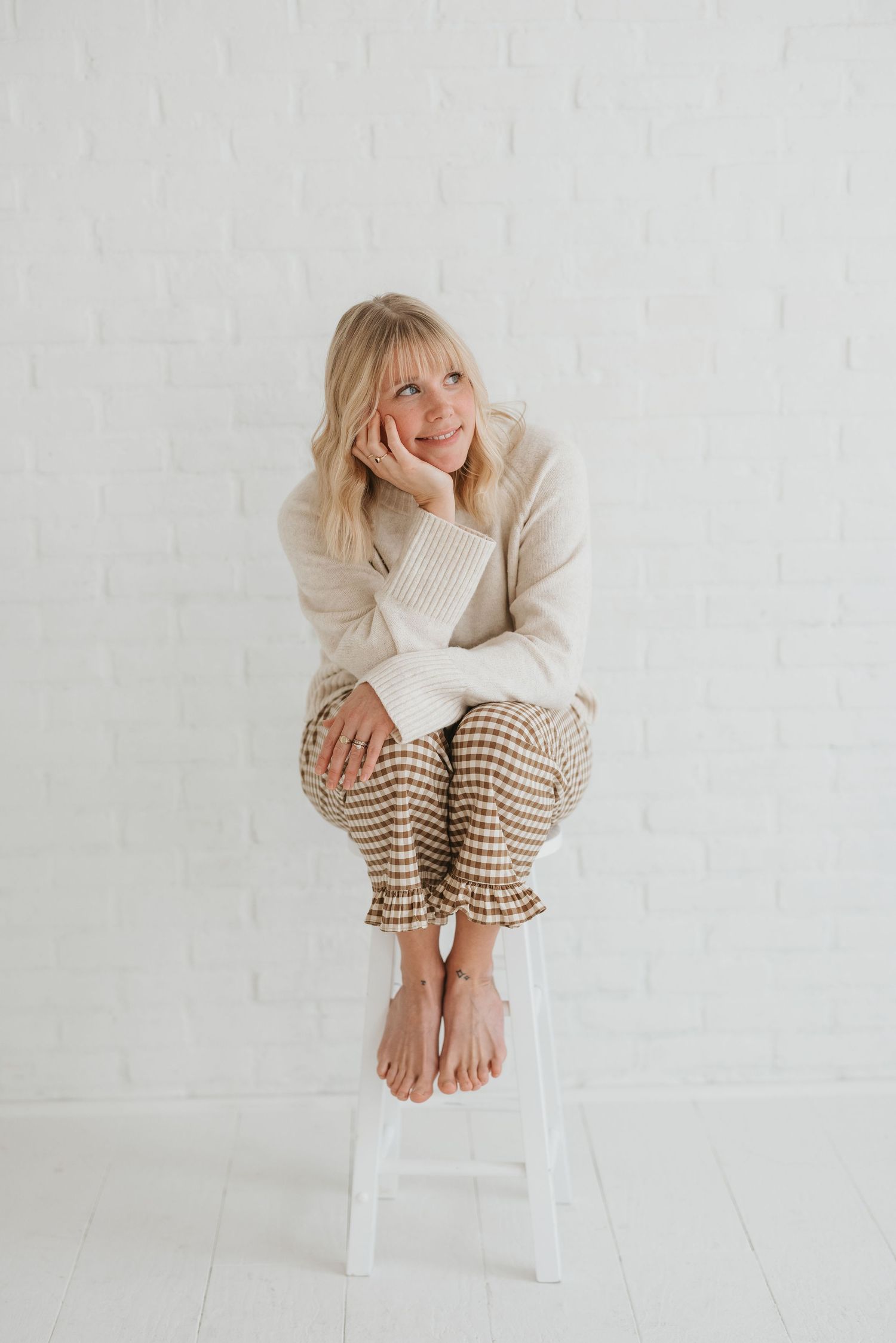 Woman in cream sweater and gingham pants dancing joyfully in bright white minimalist room.