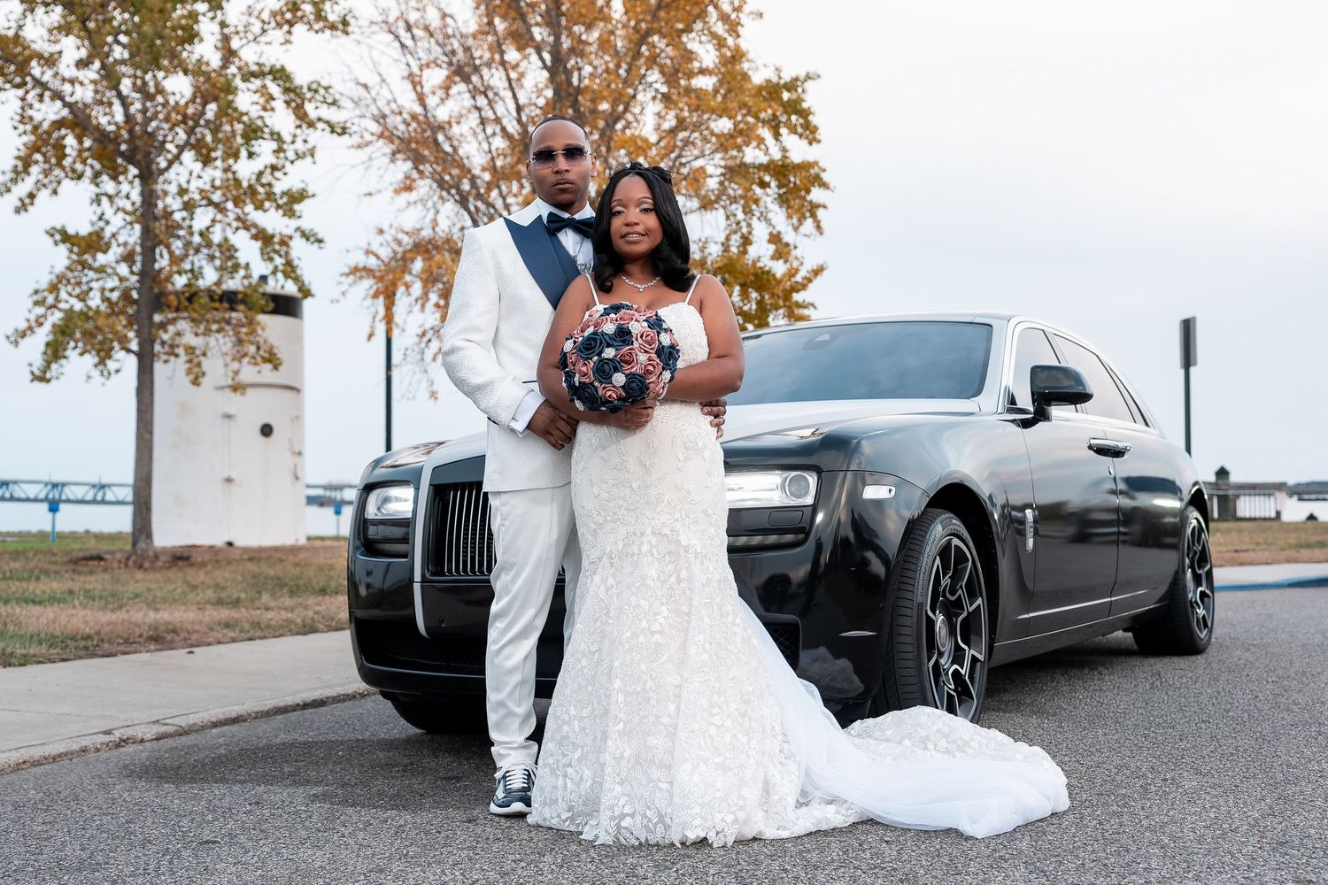 Wedding couple poses next to luxury cars in an outdoor parking lot during autumn season.