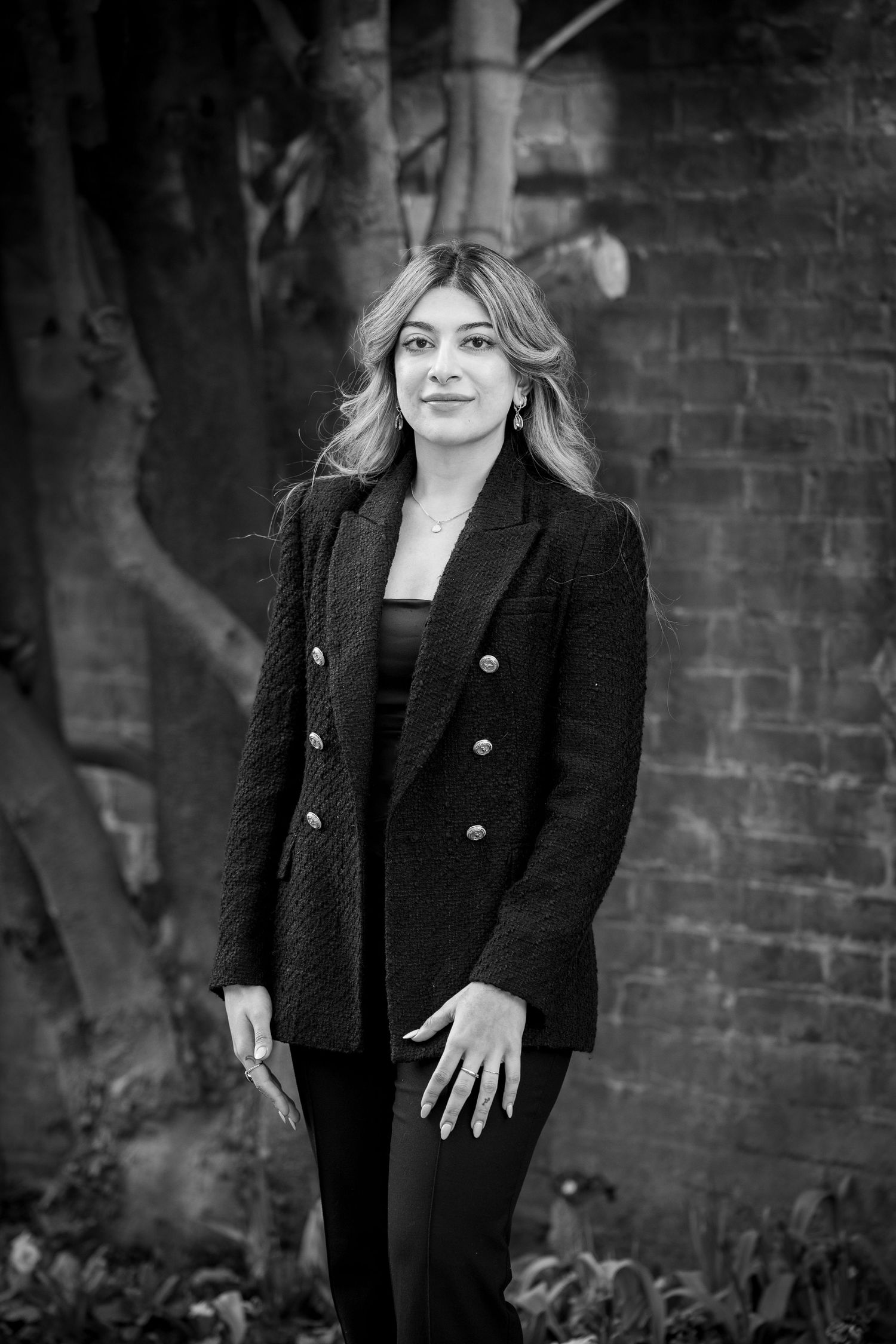 Black and white portrait of a person in a dark blazer and pants standing against a brick wall with trees in background.