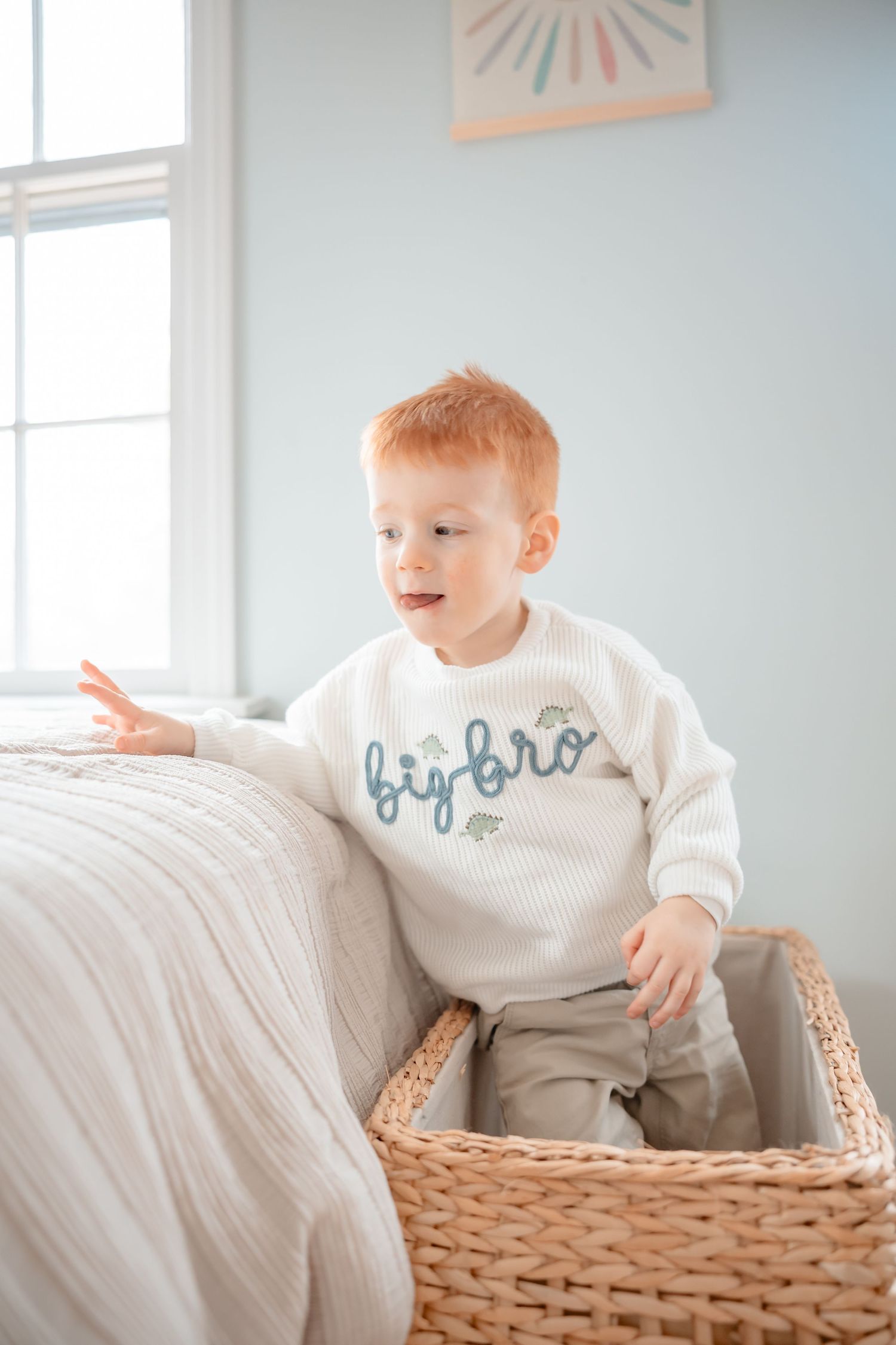 A toddler in a white sweater sits and plays in a woven storage basket against a light blue wall.