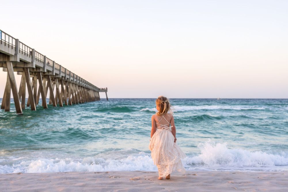 Woman in white dress standing on sandy beach beside a long pier at sunset with turquoise ocean waves.