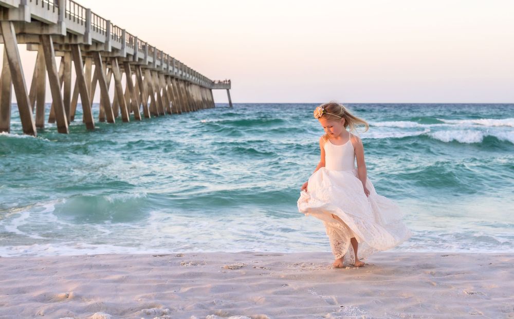 Bride in white wedding dress walks along sandy beach near wooden pier with ocean waves at sunset.