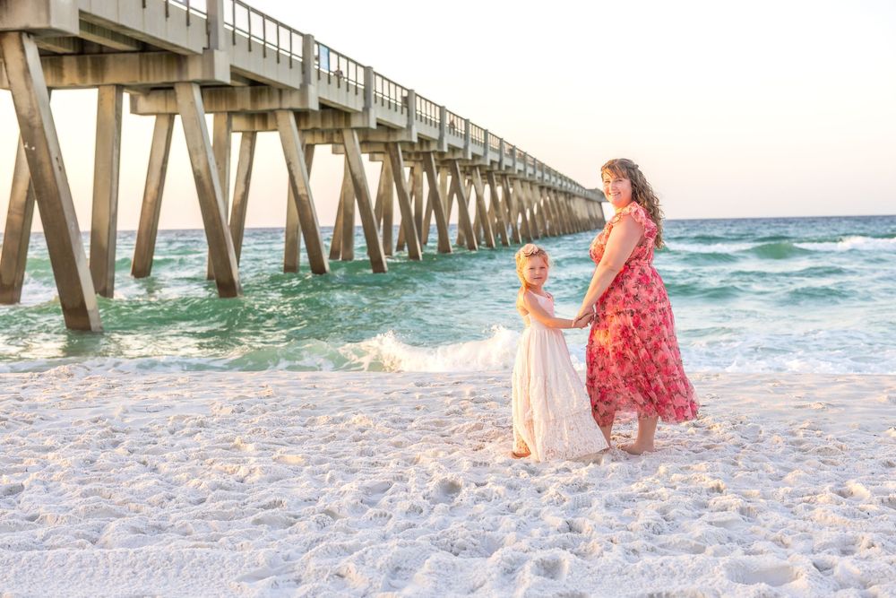 Mother and young daughter holding hands on white sand beach near a pier with turquoise ocean waves.