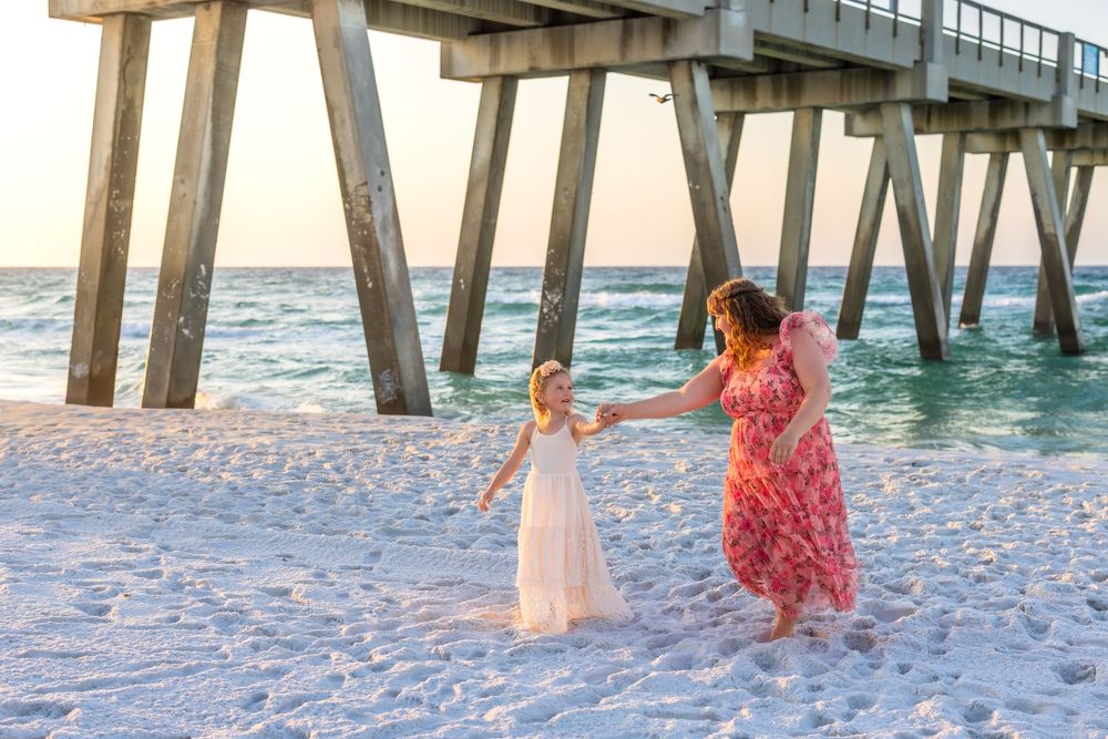 Mother and daughter dancing on white sand beach near pier at sunset in matching pink and white dresses.