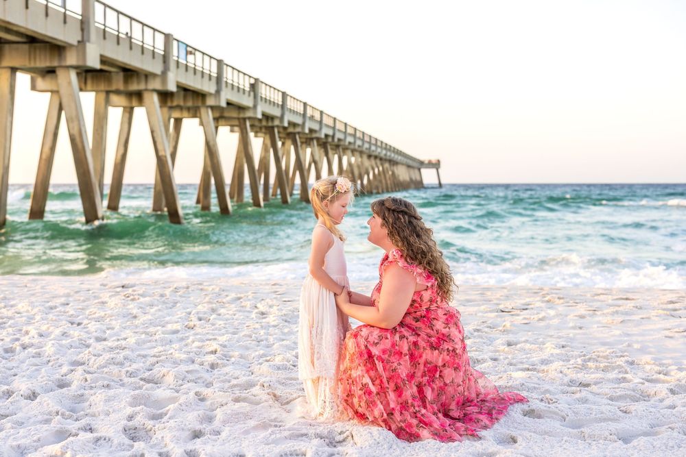 Mother and daughter in matching dresses share a tender moment on white sand beach near a wooden pier.