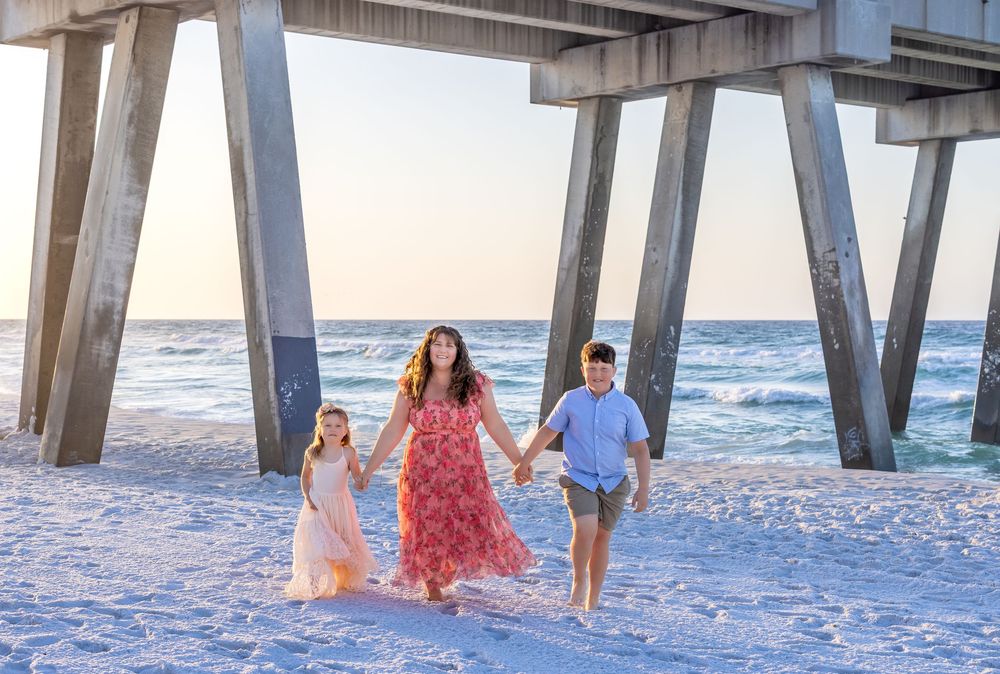 A mother and two children hold hands walking on a sunny beach near a wooden pier with ocean waves in the background.