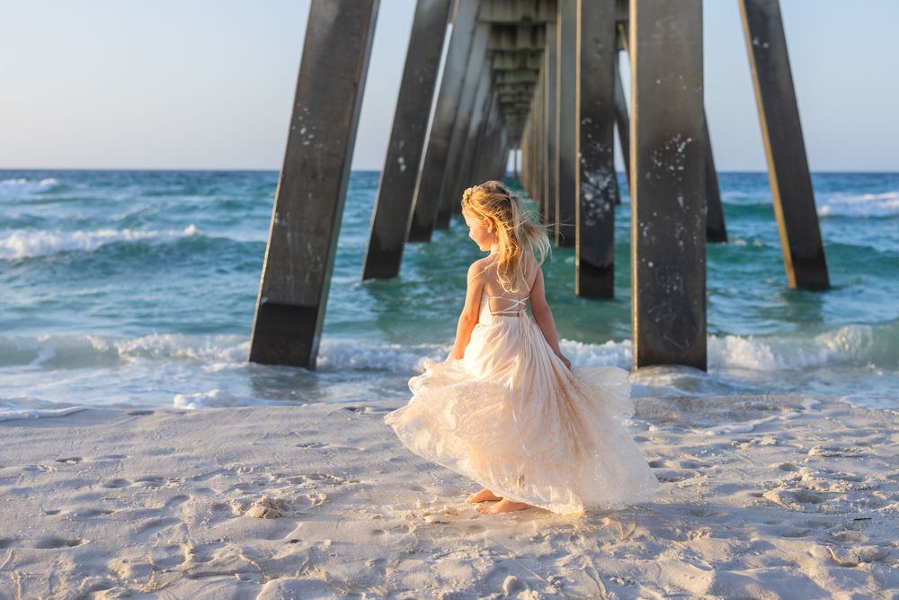 A girl in a pink ball gown twirls on a white sand beach beneath a wooden pier with ocean waves.