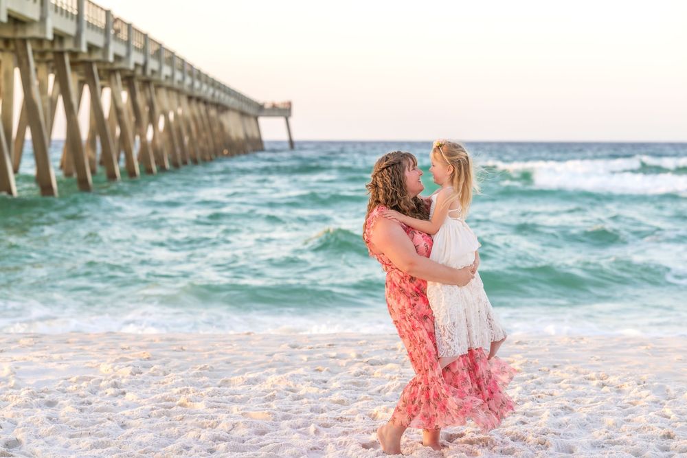 Mother and daughter dancing on a sandy beach near a pier with turquoise waves at sunset.