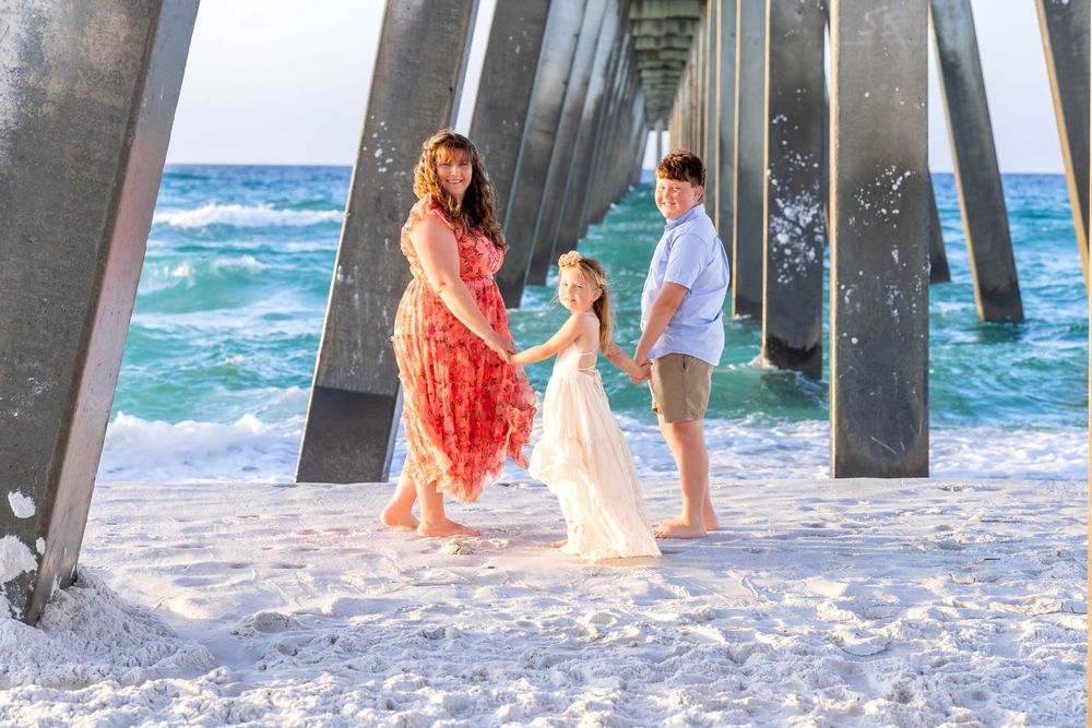 A family of three poses on a sunny beach under a wooden pier, with turquoise ocean waves behind them.