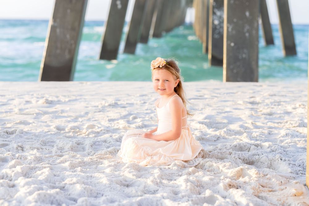 A smiling little girl in a white dress sits on white sand beach near a pier with turquoise ocean waves behind her.