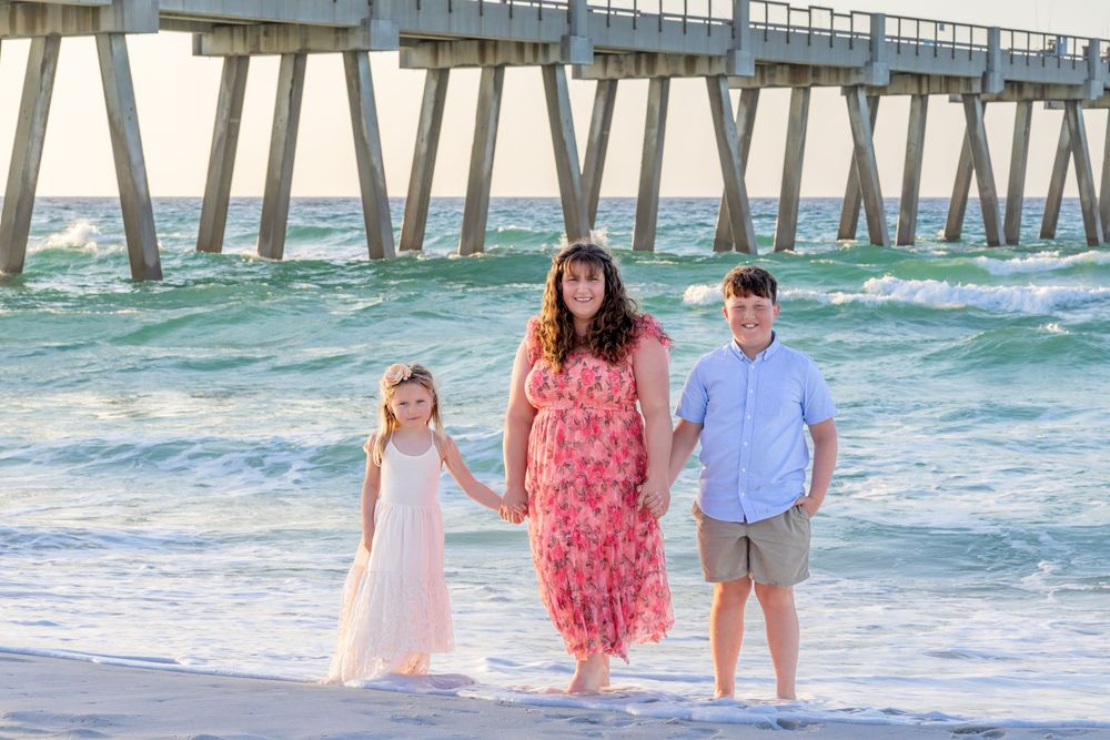 Family of three posing on a sunny beach near a pier, with turquoise waves and white sand in the background.