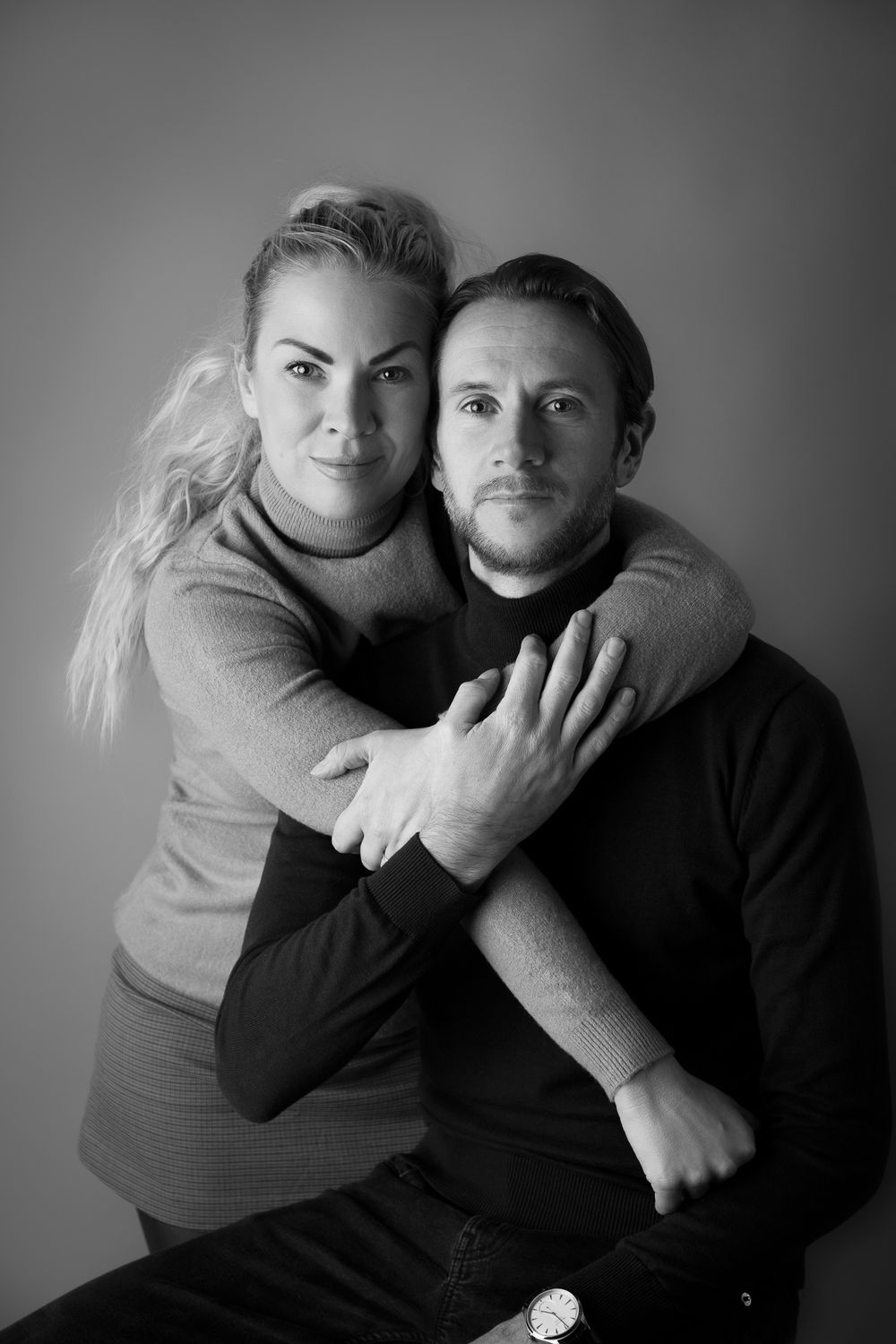 Black and white portrait of a couple, woman embracing man from behind, both looking at camera.