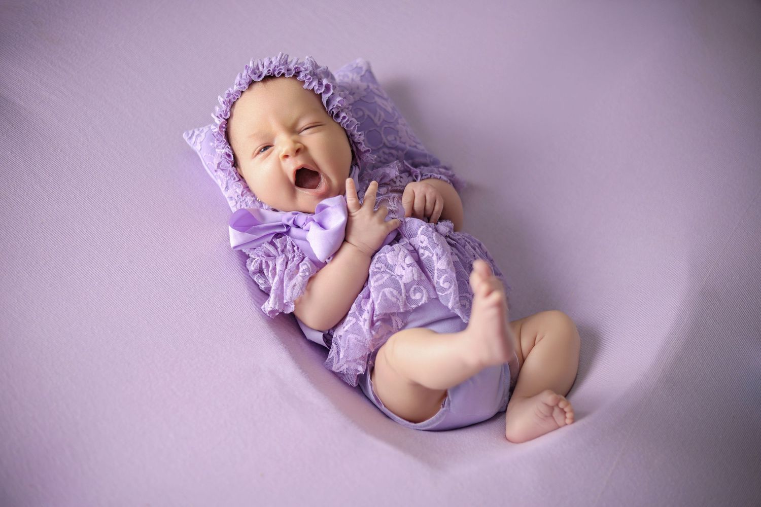 Newborn baby yawning in purple lace outfit and bonnet, lying on lavender background.