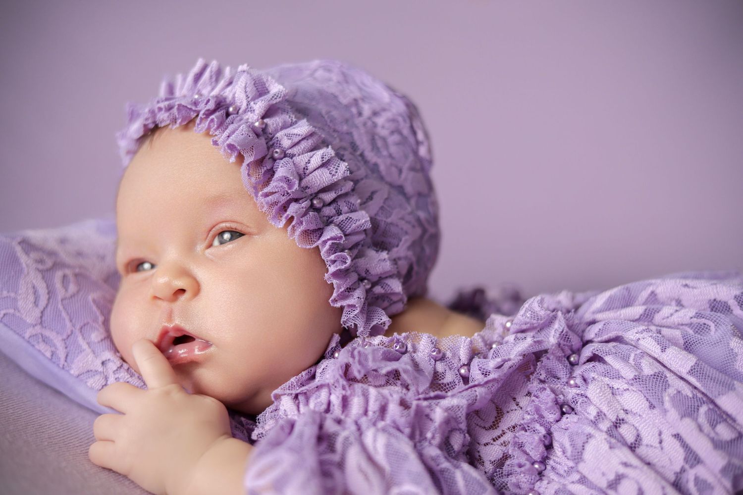 Adorable baby girl in purple lace bonnet and matching outfit lying on a soft lavender blanket.