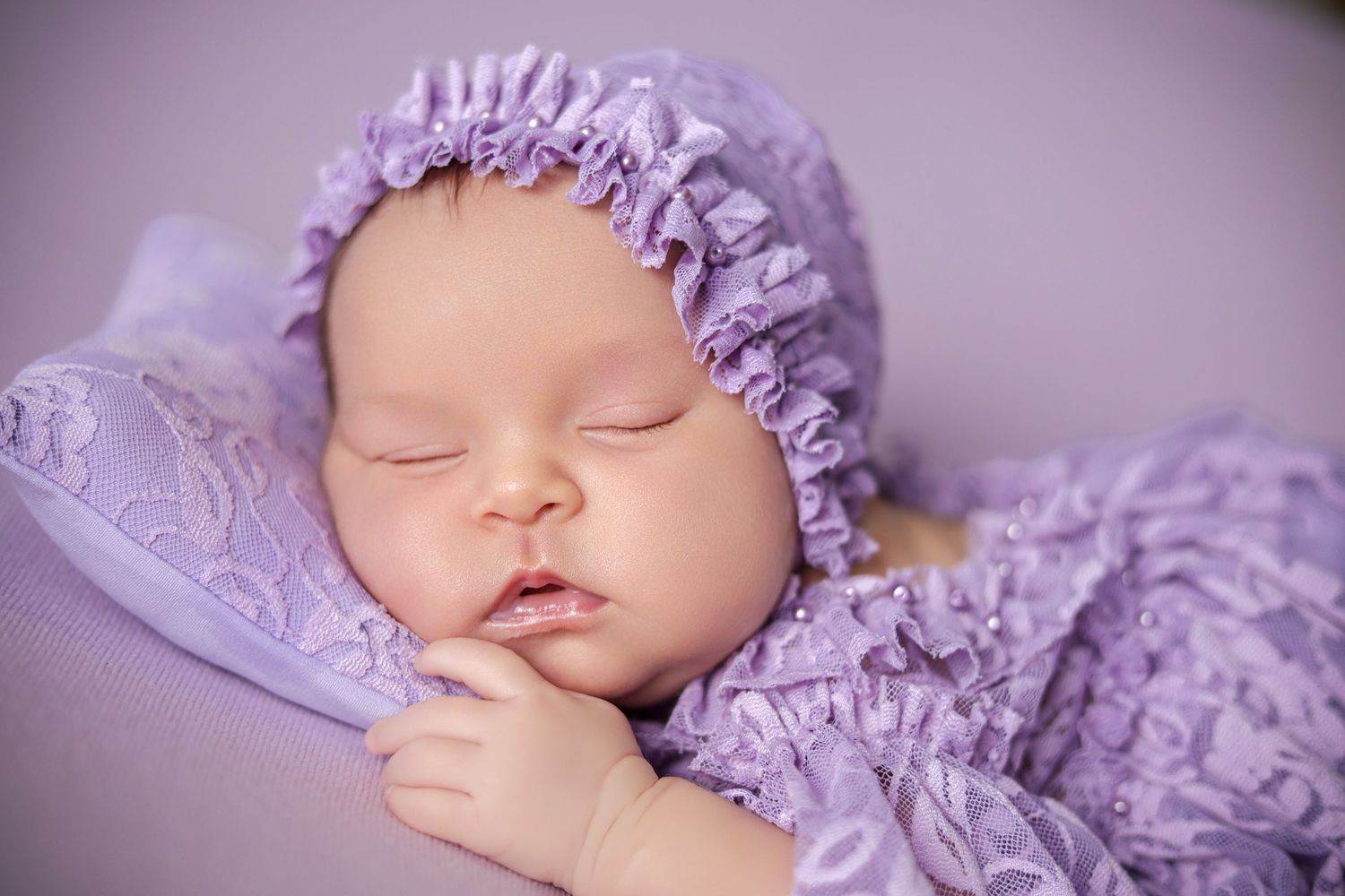 A sleeping newborn baby wearing a purple lace bonnet and matching outfit, resting peacefully on a lavender pillow.