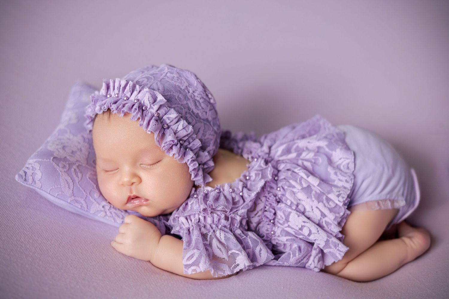 Newborn baby sleeping peacefully in purple lace outfit and bonnet on lavender background.