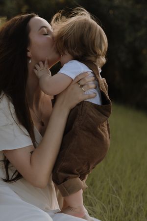 A mother in white shirt embraces and kisses her young child wearing brown in a grassy outdoor setting.