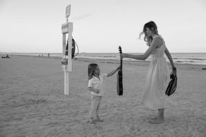 Black and white photo of people walking along a beach with a cross and guitar silhouettes at sunset.