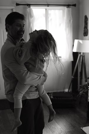 A tender black and white moment captured in a sunlit room with curtained windows and hardwood floors.
