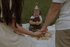 A small child in a party hat sits on a blanket outdoors while adults gather around during a celebration.