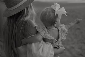A tender black and white moment shows a child in a white dress with bow being held and pointing into the distance.