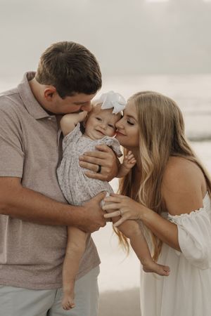 A tender family moment captured on a misty beach as parents share loving kisses with their small child.
