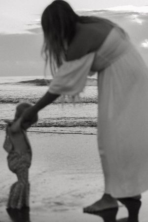 A tender black and white moment captured on a beach as a figure in white dress bends down at sunset.