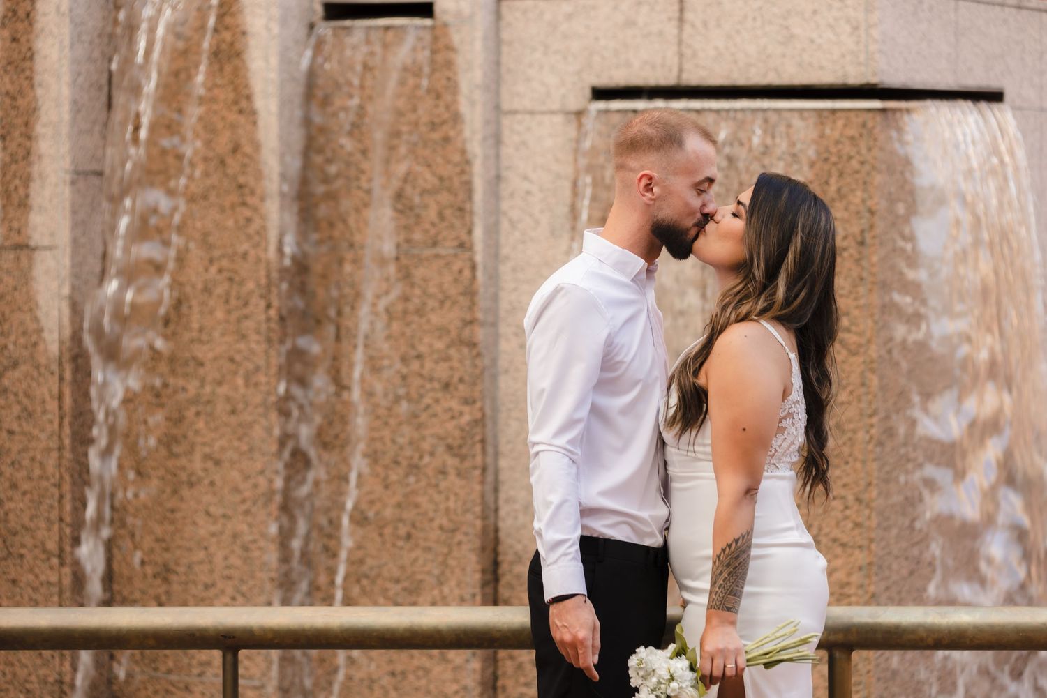 A couple in wedding attire embraces and kisses in a romantic outdoor setting with brick walls and greenery.